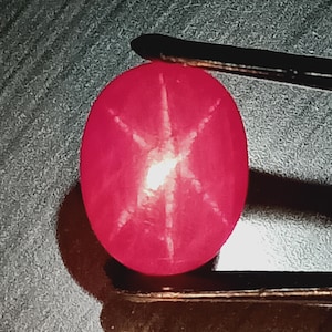 Könnte beinhalten: Ein ovalförmiger, polierter roter Edelstein mit einem sternförmigen Muster. Der Stein wird von einer Pinzette gehalten, die seine Farbe und das Sternmuster auf der Oberfläche zeigt. Die Klarheit des Edelsteins wird durch den dunklen Hintergrund hervorgehoben.