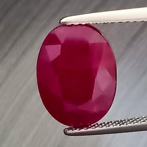 Può includere: Una gemma di rubino sfaccettata, di forma ovale, tenuta da una pinzetta. Il rubino è di un colore rosso intenso e saturo, con molteplici sfaccettature che catturano la luce. La pinzetta è color argento.