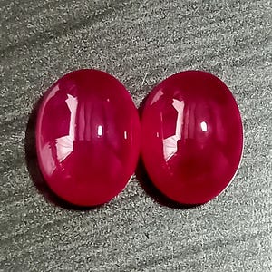 Könnte beinhalten: Zwei ovale, tief magentarote Edelsteine mit einer glatten, polierten Cabochon-Oberfläche. Jeder Stein hat eine hochglänzende Oberfläche, die das Licht reflektiert und helle weiße Glanzlichter auf den abgerundeten, roten Oberseiten erzeugt.