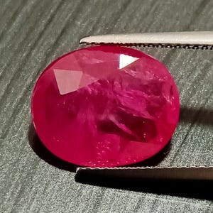 May include: A vibrant, deep red oval-shaped gemstone with a faceted cut. The gemstone is held by a pair of tweezers.