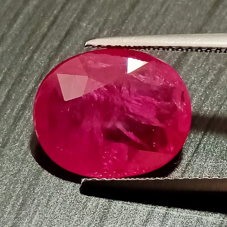 May include: A vibrant, deep red oval-shaped gemstone with a faceted cut. The gemstone is held by a pair of tweezers.