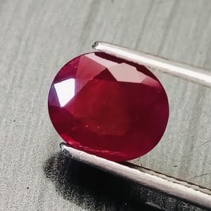 Könnte beinhalten: Ein tiefroter, oval geschliffener Edelstein mit zahlreichen Facetten, die das Licht reflektieren. Der Stein wird von einer silberfarbenen Metallpinzette gehalten und zeigt seine leuchtend karmesinrote Farbe sowie eine polierte Oberfläche.