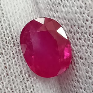May include: A vibrant, oval-cut ruby gemstone with a deep, saturated red color and multiple polished facets that reflect light. The gemstone displays a clear, transparent quality with visible internal structure and sharp, geometric edges.