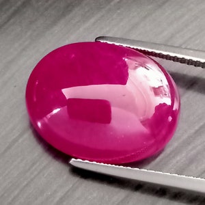 May include: A vibrant, deep pink oval-shaped gemstone with a smooth, polished surface. The gemstone is held by a pair of silver tweezers.