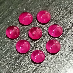 Peut inclure: Huit pierres précieuses rondes de couleur rose avec une finition lisse et polie. Les pierres précieuses sont disposées en forme de cœur sur une surface gris foncé.