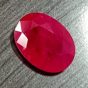 May include: A vibrant, oval-cut gemstone with a deep red color and multiple facets that reflect light. The stone features a smooth, polished surface with sharp edges along the top, showcasing a translucent quality and a rich, saturated crimson hue.