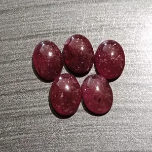 May include: Five oval-shaped, polished gemstones in a deep red hue. The gems have a smooth, reflective surface, with subtle internal inclusions. The stones are arranged on a gray surface, showcasing their color and shape.