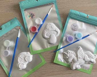 May include: Three paint-your-own kits in clear plastic bags with teal green borders. Each kit includes a white plaster figure, paint pots, and a blue paintbrush. Figures include a mushroom, car, and winged unicorn.