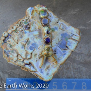 May include: A ceramic dish with a blue, green, and brown glaze. The dish has a unique, organic shape and a raised handle with colorful glass beads. The dish is approximately 5 inches wide.