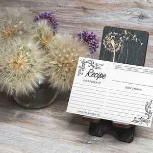 Pu&ograve; includere: Un cartoncino per ricette vuoto con un bordo decorativo e la parola "Ricetta" in corsivo. Il cartoncino &egrave; su un supporto in legno con un disegno di dente di leone.