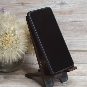 Pu&ograve; includere: Uno smartphone nero &egrave; appoggiato su un supporto in legno su una superficie in legno chiaro. Il supporto &egrave; marrone scuro con un design decorativo. Uno dente di leone bianco &egrave; sullo sfondo.