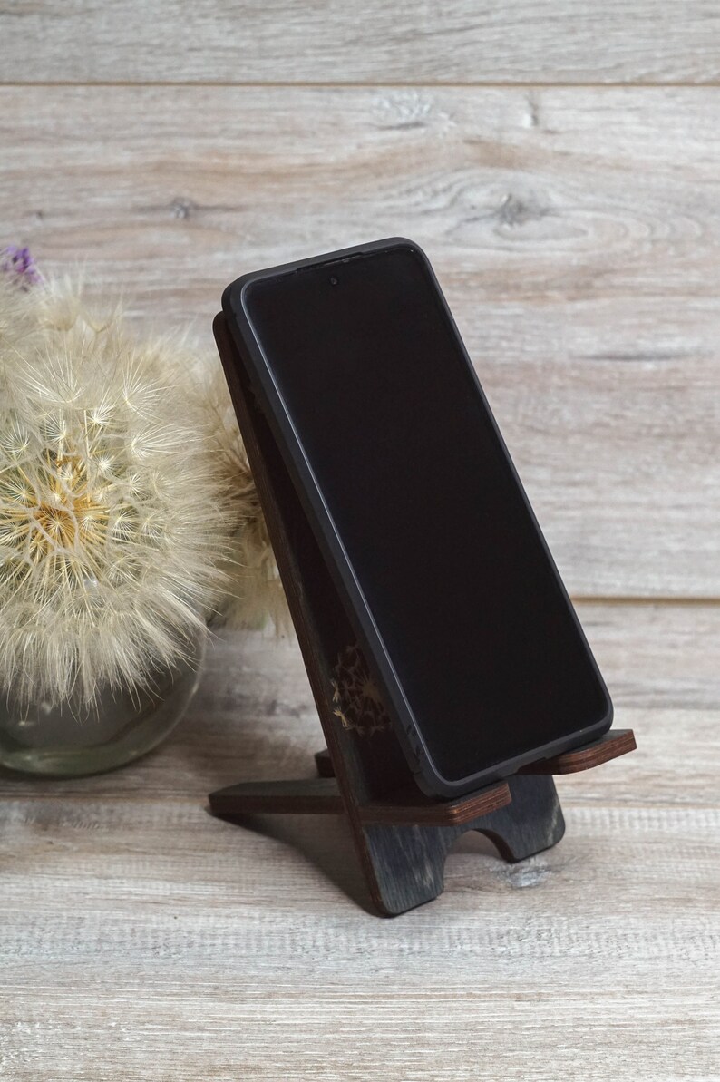 Pu&ograve; includere: Uno smartphone nero &egrave; appoggiato su un supporto in legno su una superficie in legno chiaro. Il supporto &egrave; marrone scuro con un design decorativo. Uno dente di leone bianco &egrave; sullo sfondo.