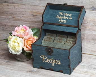 May include: A blue wooden recipe box with a hinged lid and a metal latch. The lid has the text "The Secret Ingredient Is Love" engraved on it. The box has 10 compartments for recipe cards, each with a label for different categories of recipes, such as "Drinks", "Meats", "Soups", "Salads", "Desserts", "Fish & Seafood", and "Pasta & Grains". The front of the box has the word "Recipes" engraved on it.