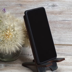 Pu&ograve; includere: Uno smartphone nero &egrave; appoggiato su un supporto per telefono in legno scuro. Il supporto ha un design semplice con due gambe e una parte superiore piatta. Il telefono &egrave; in modalit&agrave; verticale e lo schermo &egrave; nero.