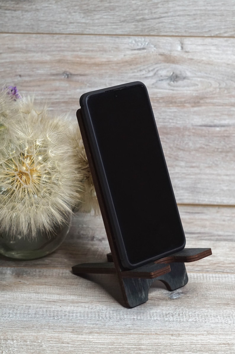 Pu&ograve; includere: Uno smartphone nero &egrave; appoggiato su un supporto per telefono in legno scuro. Il supporto ha un design semplice con due gambe e una parte superiore piatta. Il telefono &egrave; in modalit&agrave; verticale e lo schermo &egrave; nero.