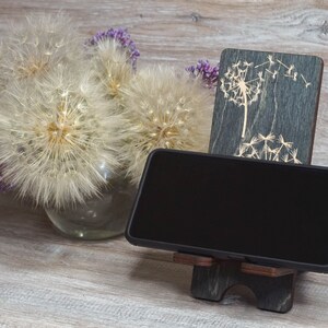 Pu&ograve; includere: Un supporto per telefono in legno con un design a dente di leone. Il supporto &egrave; grigio scuro con un design a dente di leone grigio chiaro. Uno smartphone nero &egrave; nel supporto.