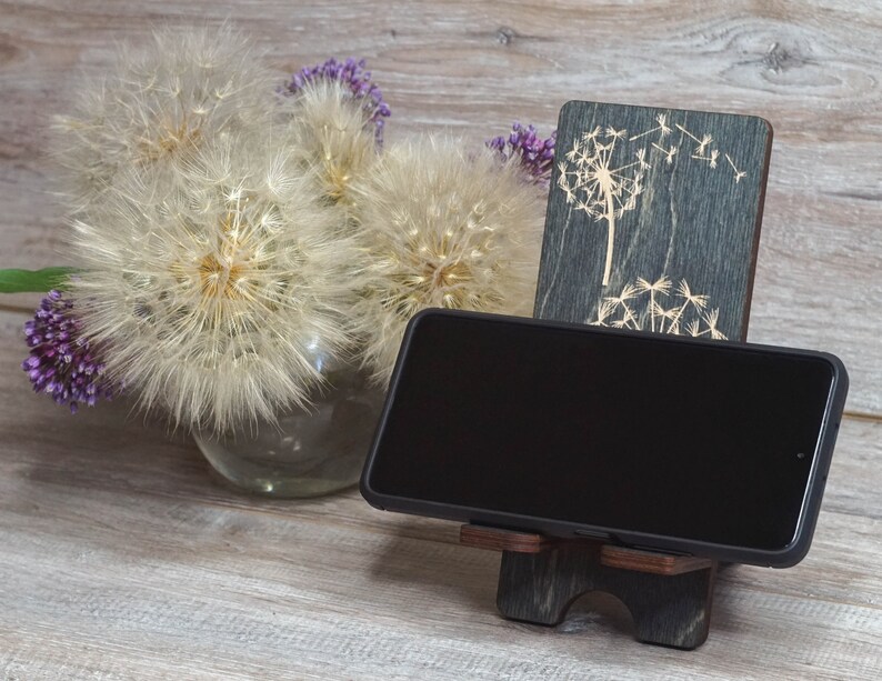 Pu&ograve; includere: Un supporto per telefono in legno con un design a dente di leone. Il supporto &egrave; grigio scuro con un design a dente di leone grigio chiaro. Uno smartphone nero &egrave; nel supporto.