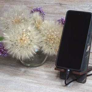 Pu&ograve; includere: Uno smartphone nero &egrave; in un supporto per telefono in legno con un cavo di ricarica collegato. Il supporto &egrave; su una superficie in legno marrone chiaro. Un vaso con fiori bianchi simili a dente di leone e fiori viola &egrave; sullo sfondo.