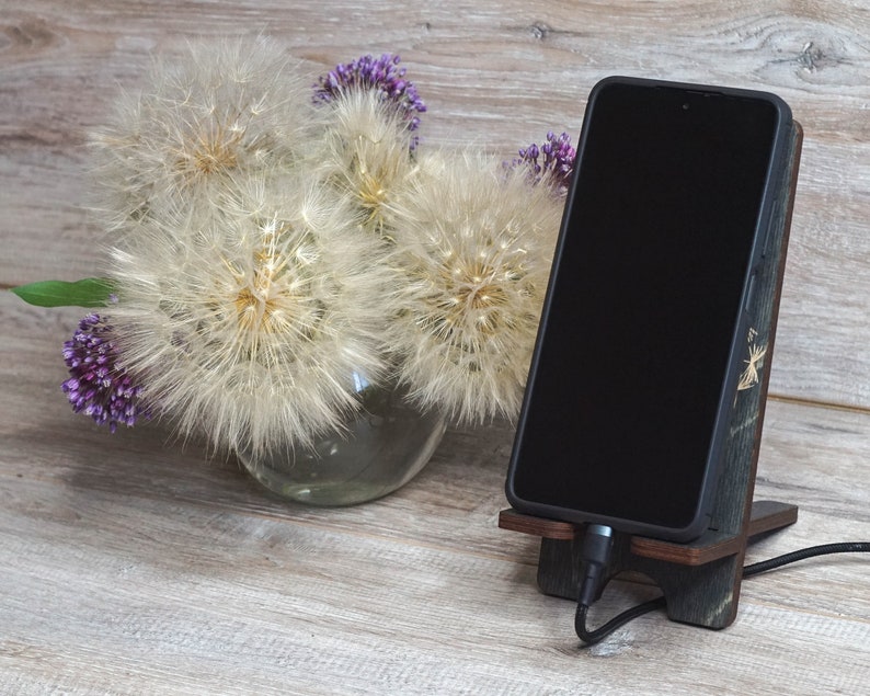 Pu&ograve; includere: Uno smartphone nero &egrave; in un supporto per telefono in legno con un cavo di ricarica collegato. Il supporto &egrave; su una superficie in legno marrone chiaro. Un vaso con fiori bianchi simili a dente di leone e fiori viola &egrave; sullo sfondo.