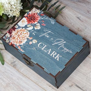 May include: A rectangular wooden keepsake box with a dark blue-grey finish and a metal latch. The lid features a printed floral design in shades of red, orange, and white, with the white text "Jim + Maggie CLARK" printed on the surface.