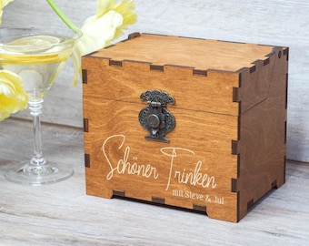 May include: A wooden box with a hinged lid and a metal clasp. The box is engraved with the text "Schöner Trinken mit Steve & Jul".