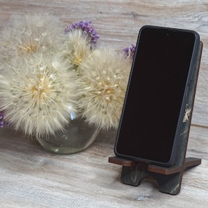 Pu&ograve; includere: Un supporto per telefono in legno con finitura in legno scuro e un accento in legno chiaro. Il supporto &egrave; decorato con un piccolo design dorato. Uno smartphone nero &egrave; appoggiato sul supporto.