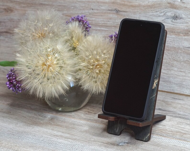 Pu&ograve; includere: Un supporto per telefono in legno con finitura in legno scuro e un accento in legno chiaro. Il supporto &egrave; decorato con un piccolo design dorato. Uno smartphone nero &egrave; appoggiato sul supporto.