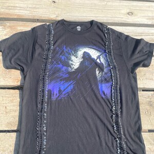 May include: Black t-shirt featuring a graphic of the Grim Reaper with a scythe, set against a blue and white background. The shirt is embellished with rows of silver safety pins along the sides, adding a unique, edgy design.