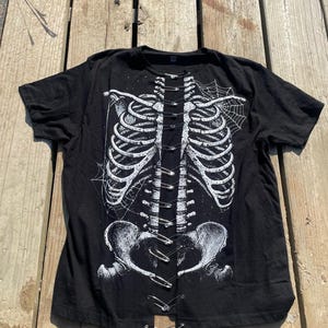 Könnte beinhalten: Schwarzes T-Shirt mit einem weißen Skelett-Brustkorb-Design und Spinnweben. Der Brustkorb wird mit Sicherheitsnadeln zusammengehalten. Das T-Shirt ist aus einem weichen Material.
