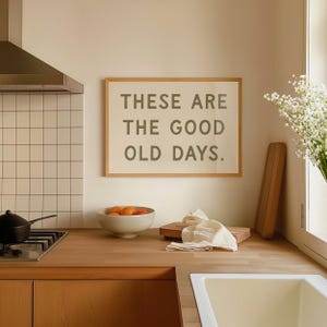 May include: A framed print with the text "These are the good old days." hanging on a light beige wall in a kitchen. The print is in a light wood frame. There is a bowl of oranges on the counter below the print.