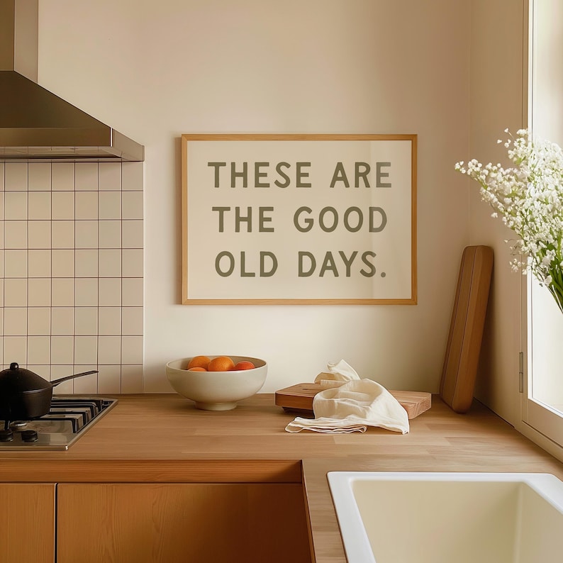 May include: A framed print with the text "These are the good old days." hanging on a light beige wall in a kitchen. The print is in a light wood frame. There is a bowl of oranges on the counter below the print.