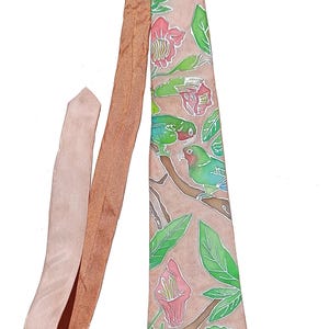 Pode incluir: Uma gravata num tom de rosa poeirento suave com um design pintado à mão de dois papagaios verdes pousados em ramos castanhos. O padrão inclui folhas verde-brilhante e flores cor-de-rosa com contornos brancos sobre o tecido rosa sólido.