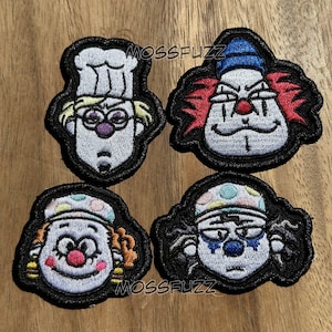 Puede incluir: Cuatro parches bordados con caras de payasos con diversas expresiones y detalles coloridos. Cada parche tiene un borde negro y está sobre un fondo de madera. Los parches incluyen un payaso chef, un payaso con pelo rojo y otros dos diseños de payasos.