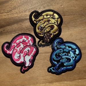 Könnte beinhalten: Drei gestickte Bügelbilder mit verschiedenen Schlangenmustern. Die Schlangen sind rosa und weiß, blau und schwarz sowie gold und braun.