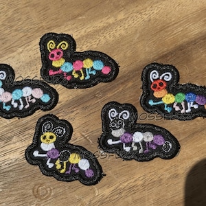 May include: Five embroidered caterpillar patches with black borders and vibrant colours. Each caterpillar features a unique colour palette, including blue, pink, yellow, and purple. The text "MOSSFUZZ" is visible on the wooden surface.