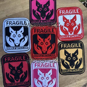 May include: A selection of square patches, each displaying the word "FRAGILE" at the top. The patches feature a wolf head and hands design, with the text "HANDLE WITH CARE" at the bottom. The patches are in various colour combinations.