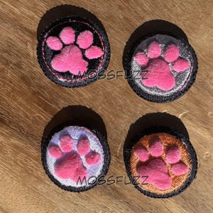 May include: Four round patches with a pink paw print design. Each patch has a black border and a different colored background: black, silver, lavender, and orange. The text "MOSSFUZZ" is printed on two of the patches.