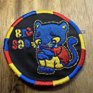 May include: Embroidered patch featuring a blue cat with yellow eyes, crying tears. The cat is wearing a red vest and yellow pants. The words "BIG SAD" are embroidered in yellow and red. The patch has a black background with a red, yellow, and blue border.