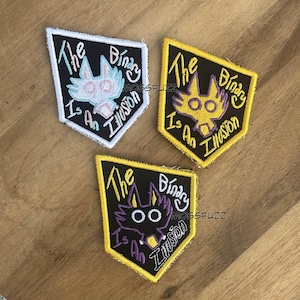 May include: Three embroidered patches with a black background and yellow border. Each patch features a cartoon animal with large eyes and the text "The Binary Is An Illusion". The animals are a white fox, a yellow fox, and a purple fox.