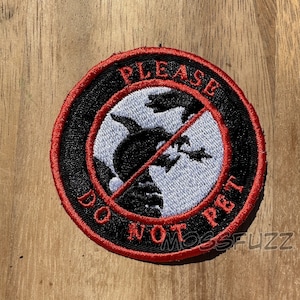 May include: Circular embroidered patch with a black and red border. The patch features a black silhouette of a creature with the words "PLEASE DO NOT PET" in red. The background is a light blue and white design.