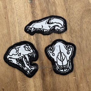 以下が含まれることがあります： 動物の頭蓋骨が刺繍された、白黒のワッペンが3つ。頭蓋骨はオオカミ、キツネ、クマのものです。ワッペンは木製の表面に置かれています。