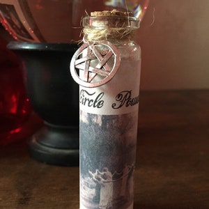 May include: A small glass bottle with a cork stopper and a silver pentagram charm. The bottle is labeled "Circle Power" and has a black and white image of a group of people holding hands in a circle.