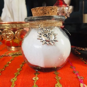 May include: A small glass jar with a cork lid and a silver sun charm. The jar is filled with a white powder and is sitting on a red and gold sequined surface.