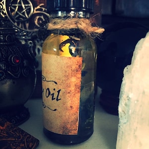 May include: A small glass bottle with a brown label that says "Oil" on it. The bottle is filled with a clear liquid and has a black cap. The bottle is sitting on a white surface.