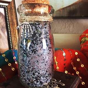 May include: A glass bottle with a cork stopper filled with black and white salt crystals. The bottle has a silver charm attached to it in the shape of a skeleton hand.