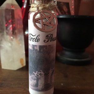 May include: A small glass bottle with a cork stopper and a silver pentagram charm. The bottle is labeled "Circle Power" and contains a white powder. The bottle is sitting on a wooden surface.