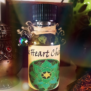 May include: A small glass bottle with a black cap and a label that says "Heart Chakra". The label has a green and blue mandala design with a yellow star in the center. The bottle is filled with a clear liquid and has a small, iridescent crystal charm attached to the cap.