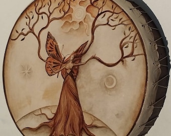 May include: Round drum with a painted design featuring a tree with a butterfly figure. The artwork is in shades of brown, beige, and orange, with a sun, moon, and star. The drum's edge is stitched with a natural-colored material.