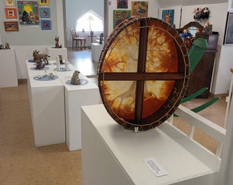 May include: A large, round, wooden drum with a brown leather head. The drum is decorated with a painted design of a tree with branches. The drum is on display in a gallery setting.