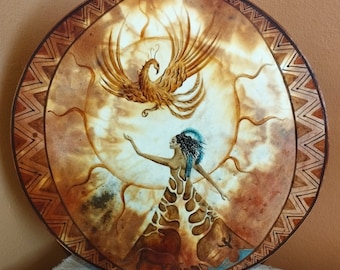 May include: A round, brown leather drum with a painted design of a woman reaching towards a golden phoenix. The phoenix is flying in a circle around a sun with rays. The woman is wearing a blue and white headdress and a brown dress with a pattern.